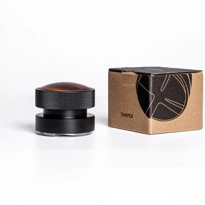 Black coffee tamper with textured grip and wooden top next to cardboard box on white background
