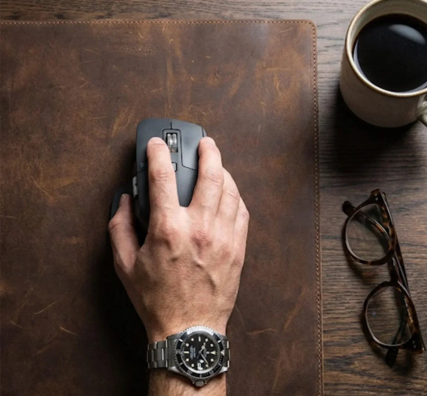 Hand using a wireless mouse on a leather desk pad with coffee and eyeglasses nearby